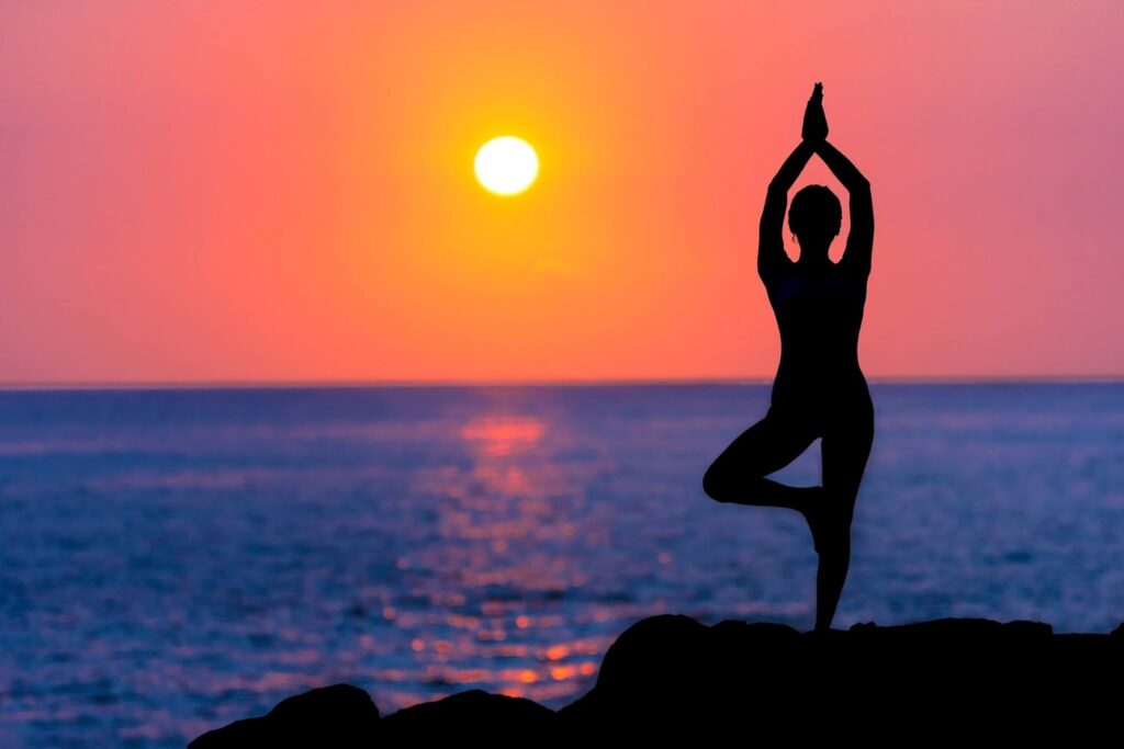 The silhouette of a woman doing yoga on the beach against the setting sun near the horizon.